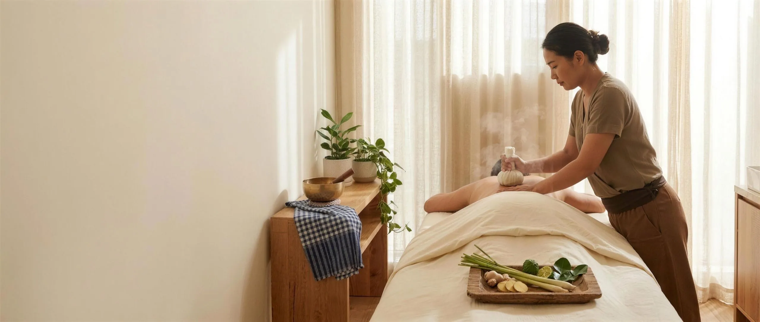 Therapist pressing a steaming Thai herbal compress on a client's back with a tray of herbs nearby